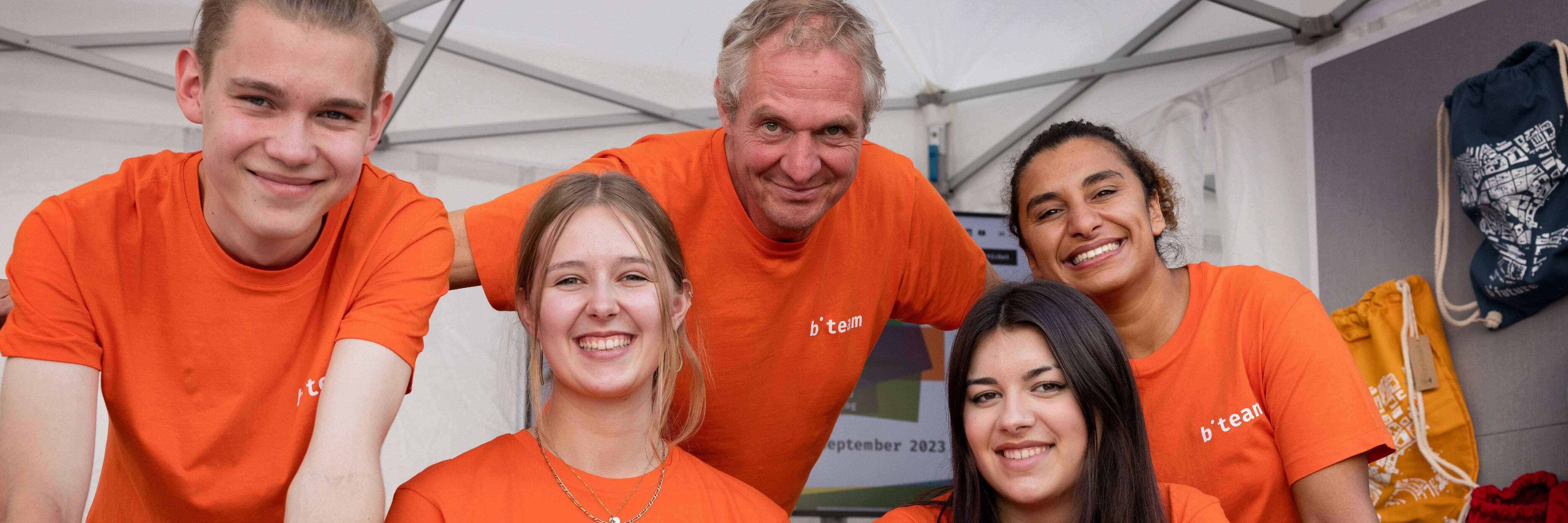 Five team members of the b-future team, dressed in orange, stand at a festival booth, smiling warmly at the camera. They are wearing orange T-shirts with the inscription "b-team."