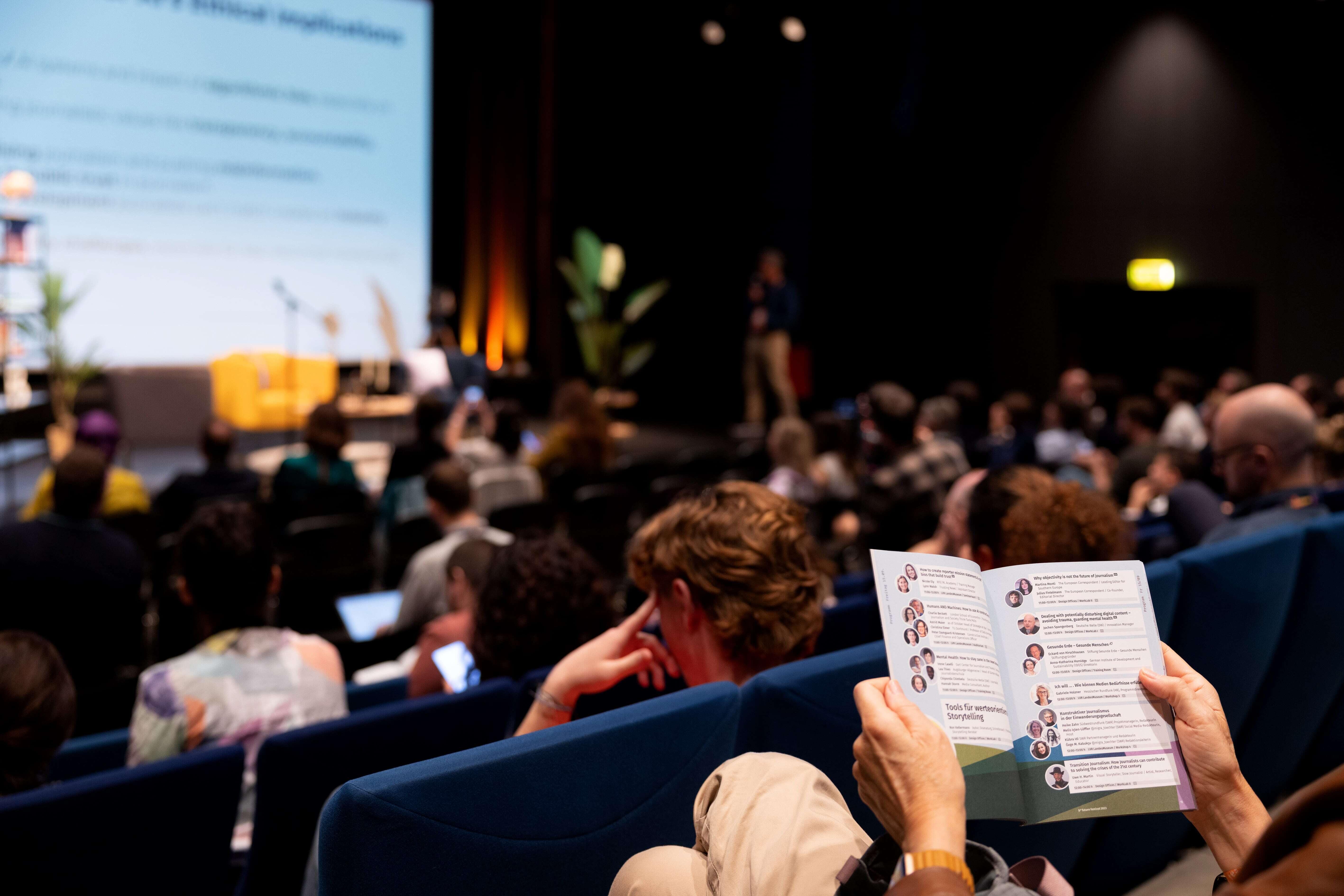 In Reihen sitzt das Publikum des Festivals auf blauen Sesseln und schaut in Richtung der Bühne. Eine Besucherin liest im aufgeschlagenen Programm des Festivals.