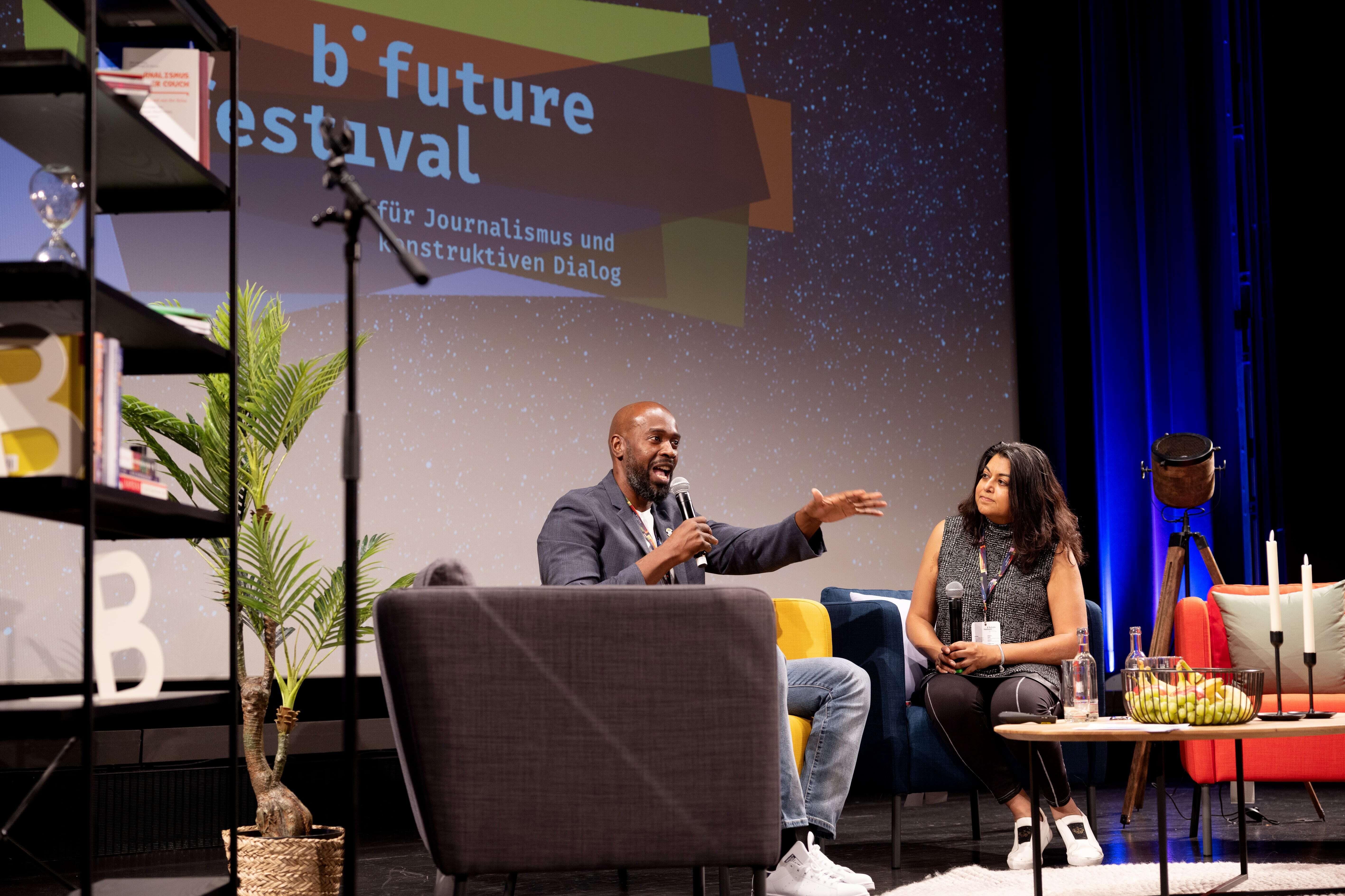 Derrick Cain and Aparna Mukherjee are speaking on the stage, in the background, you can see the colorful b° future logo on a large screen.