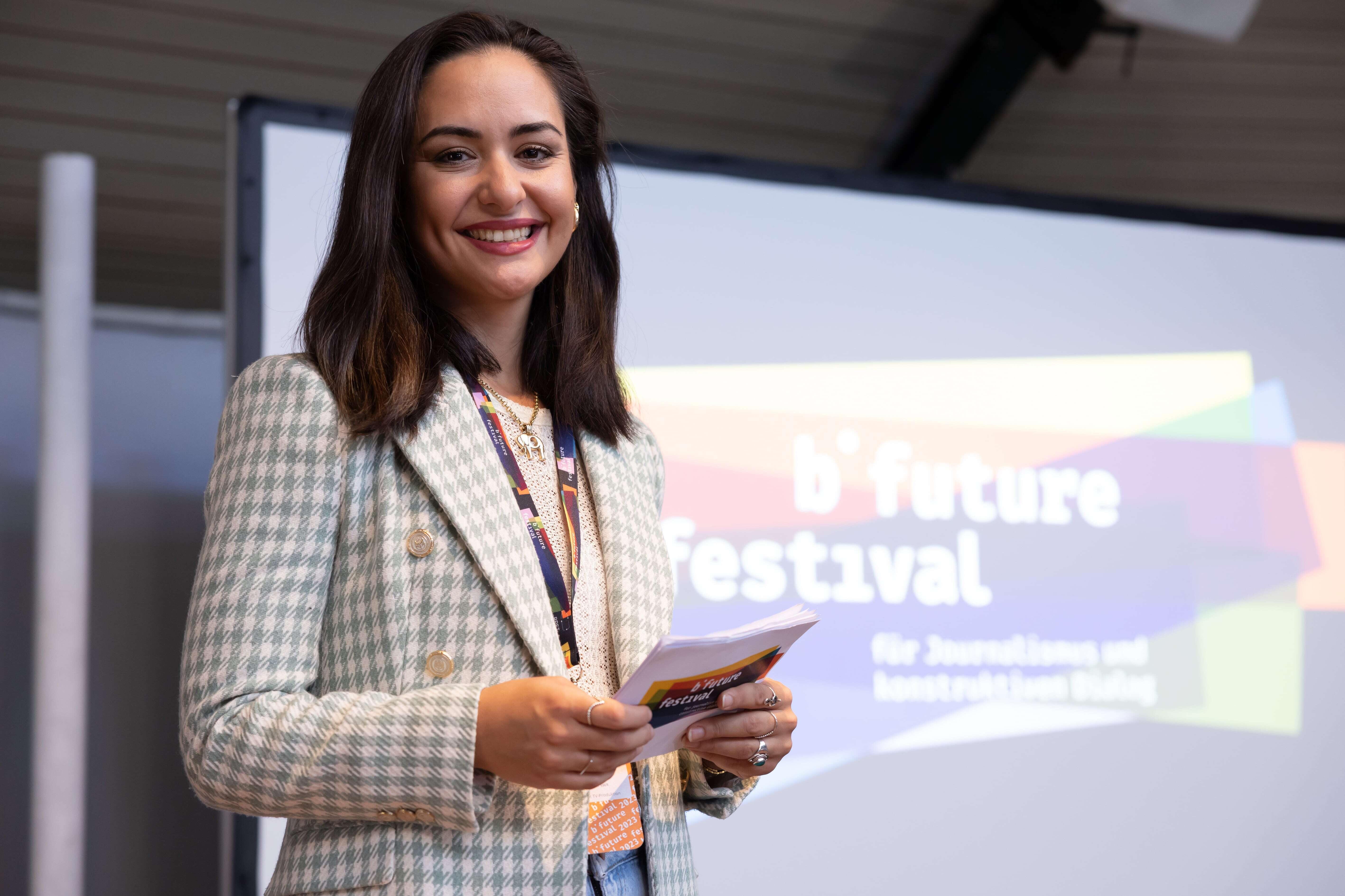 Özge M. Kabukçu lächelt in die Karte während ihrer Moderation auf der Bühne. Sie hält Moderationskarten in der Hand, hinter ihr sieht man das Logo des b° future Festivals auf einem Bildschirm.