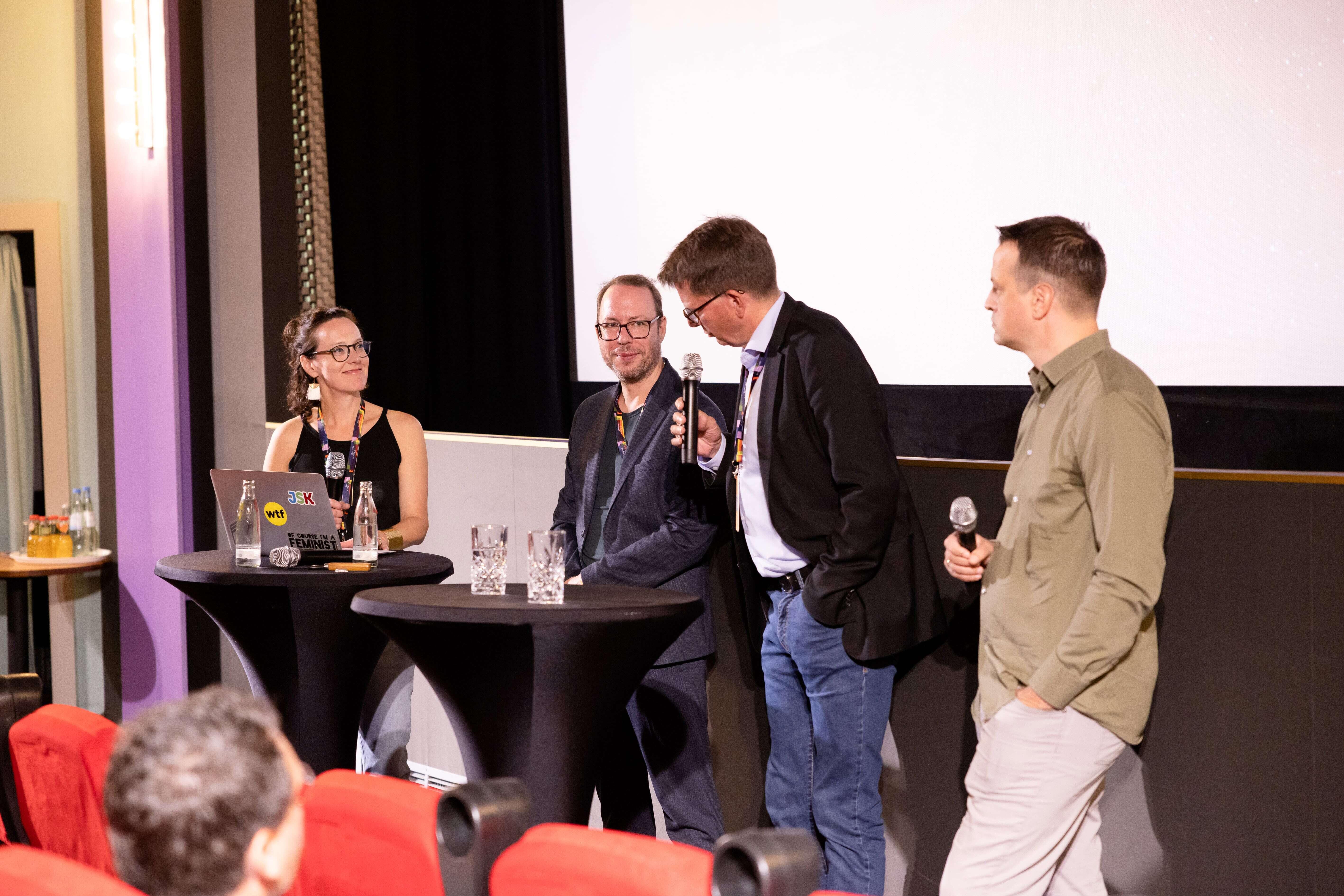 Von links nach rechts stehen Astrid Maier, Markus Beckedahl, Moritz Döbler und Simon Hof an zwei Stehtischen und sprechen in Mikrofone. Vor ihnen rote Stuhlreihen und hinter ihnen eine große weiße Leinwand.
