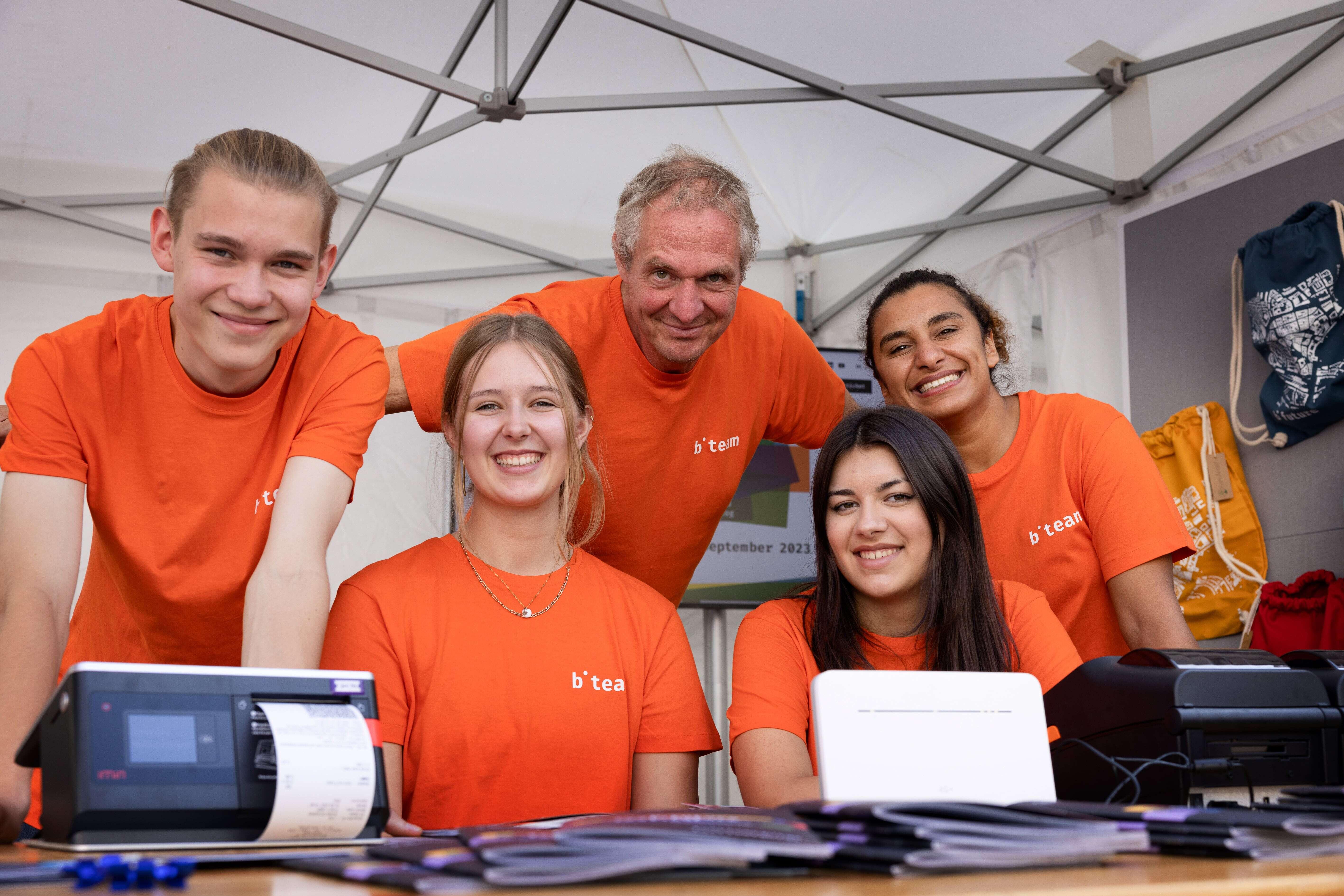 Five team members of the b-future team, dressed in orange, stand at a festival booth, smiling warmly at the camera. They are wearing orange T-shirts with the inscription "b-team."