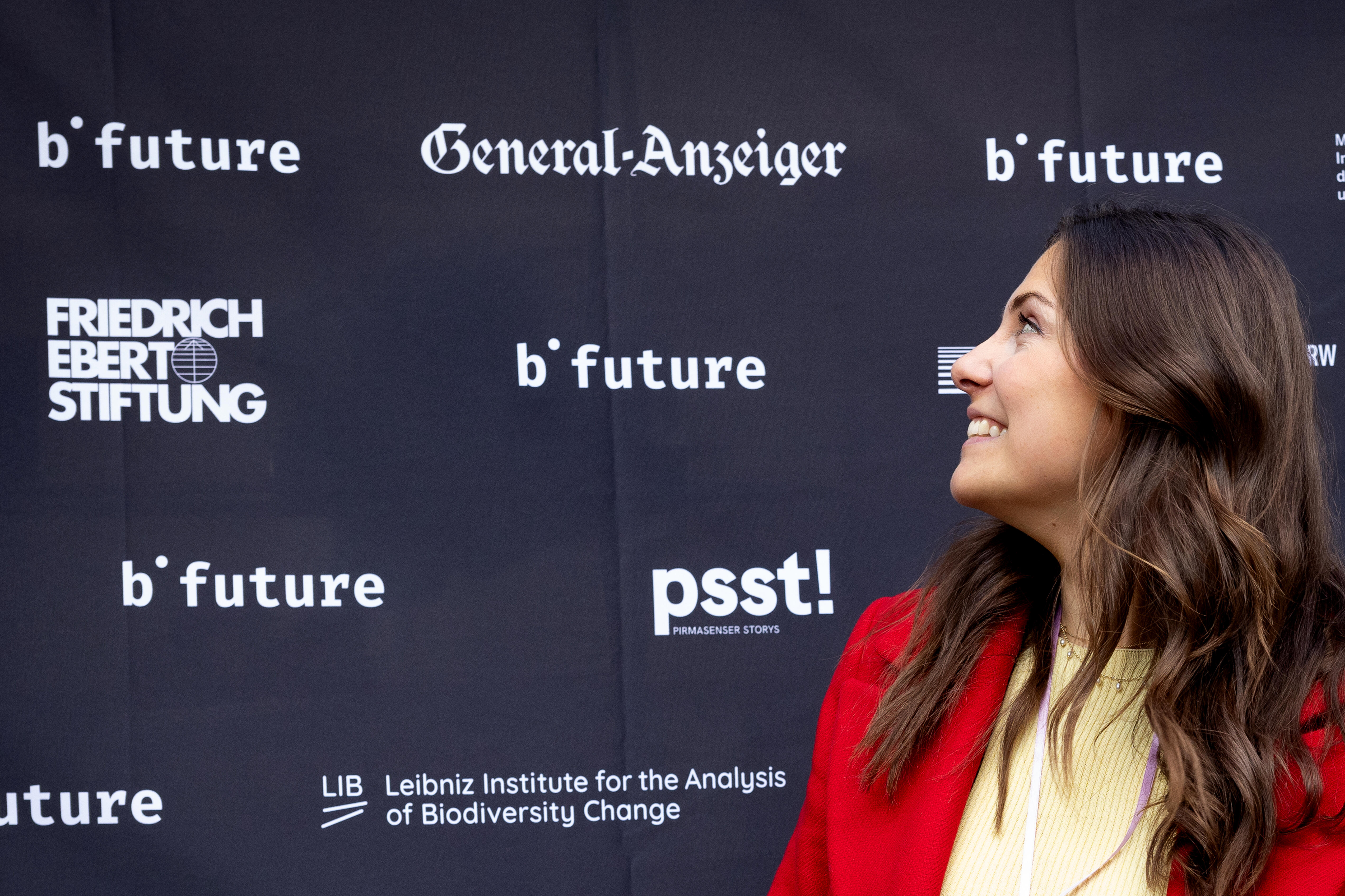 Im Hintergrund Logowand mit verschiedenen Partner des b° future festival. Vordergrund Spenderin Clara Pfeffer in einem roten Mantel