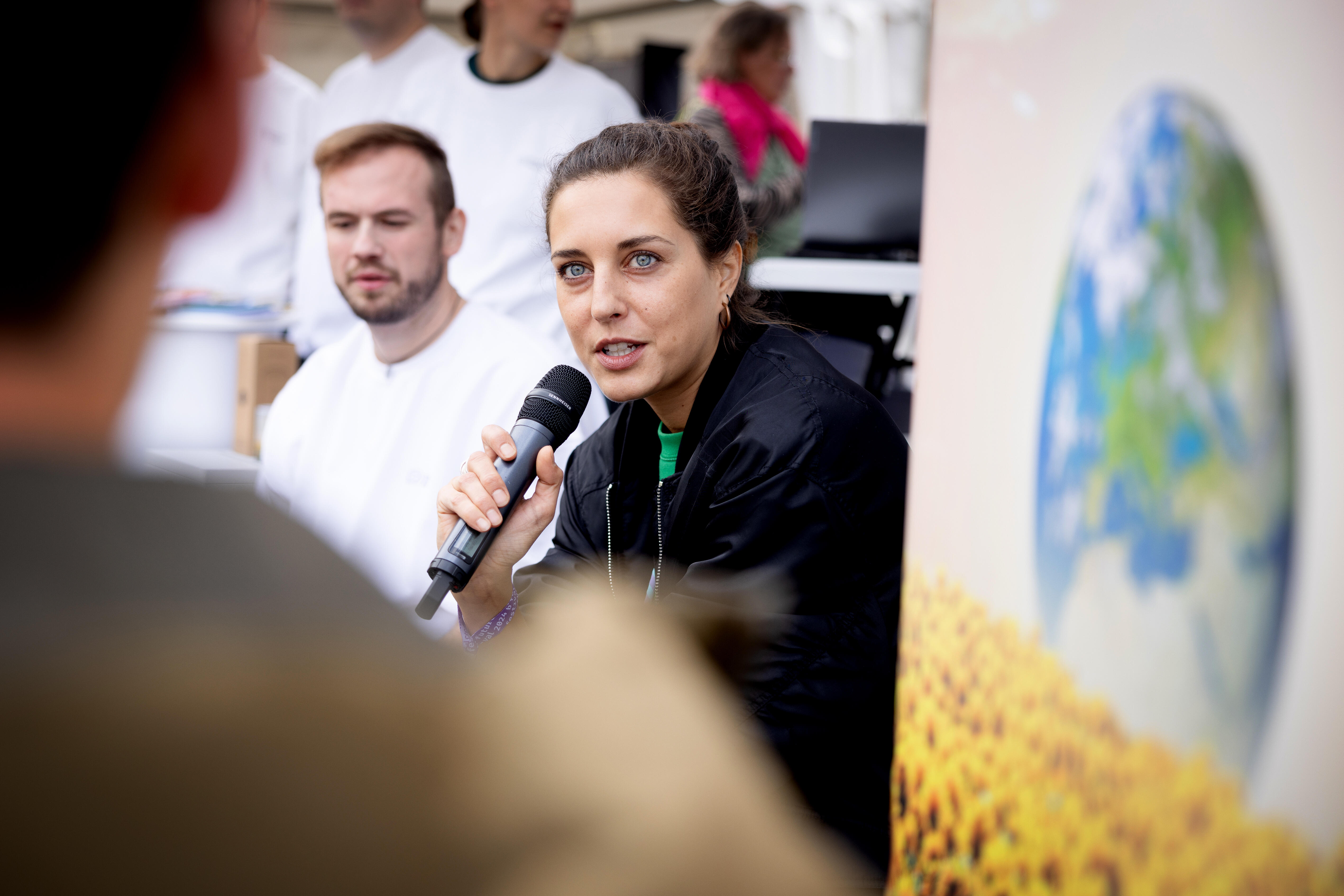 Menschen auf dem festival, im Vordergrund verschwommen Publikum welches zuhört. Zu sehen ist eine Frau mit Mikrofon in der Hand und auch im Hintergrund Publikum.