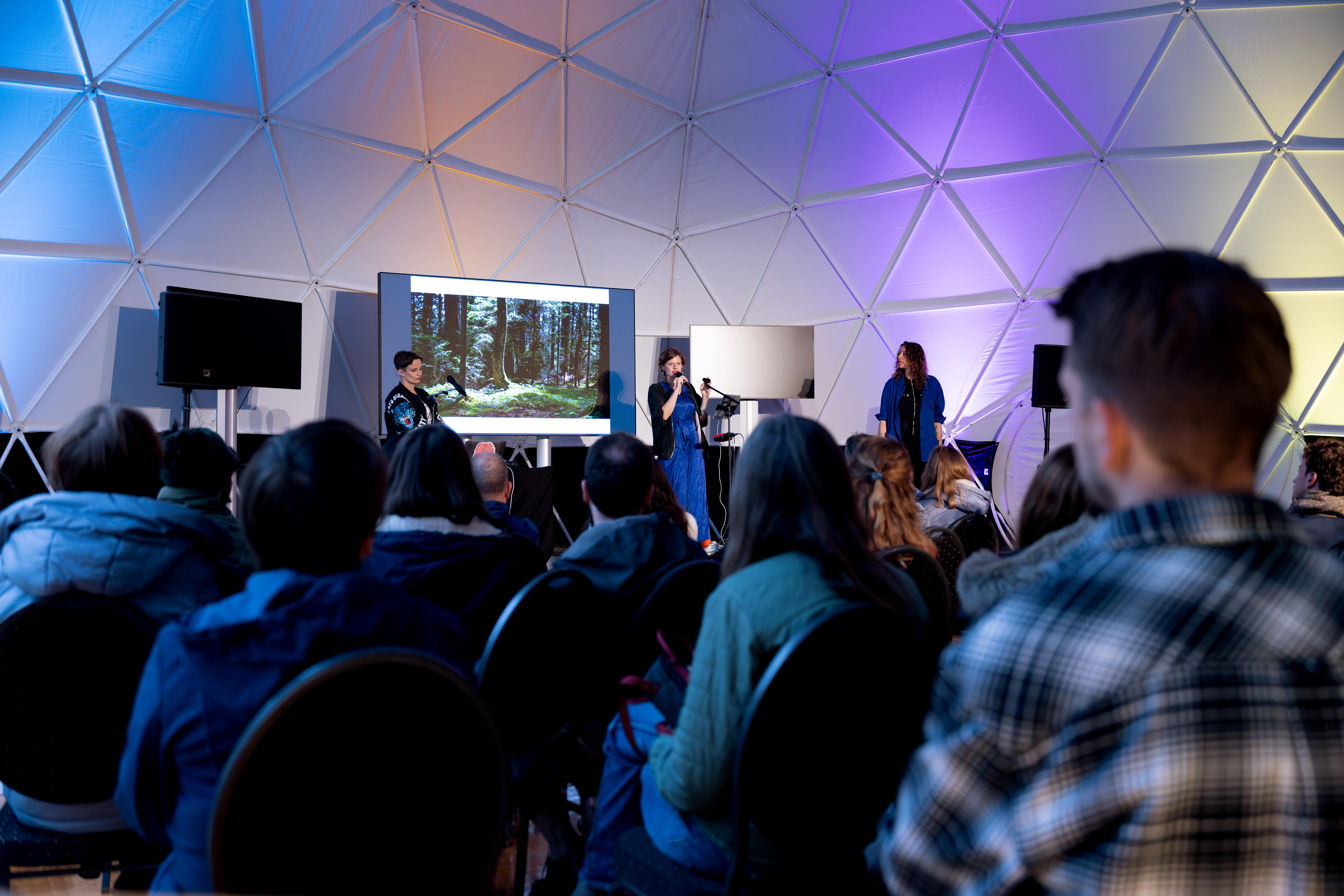 Eine Hauptbühne auf dem ein Vortrag stattfindet. Vor der Bühne sitzen viele Leute.