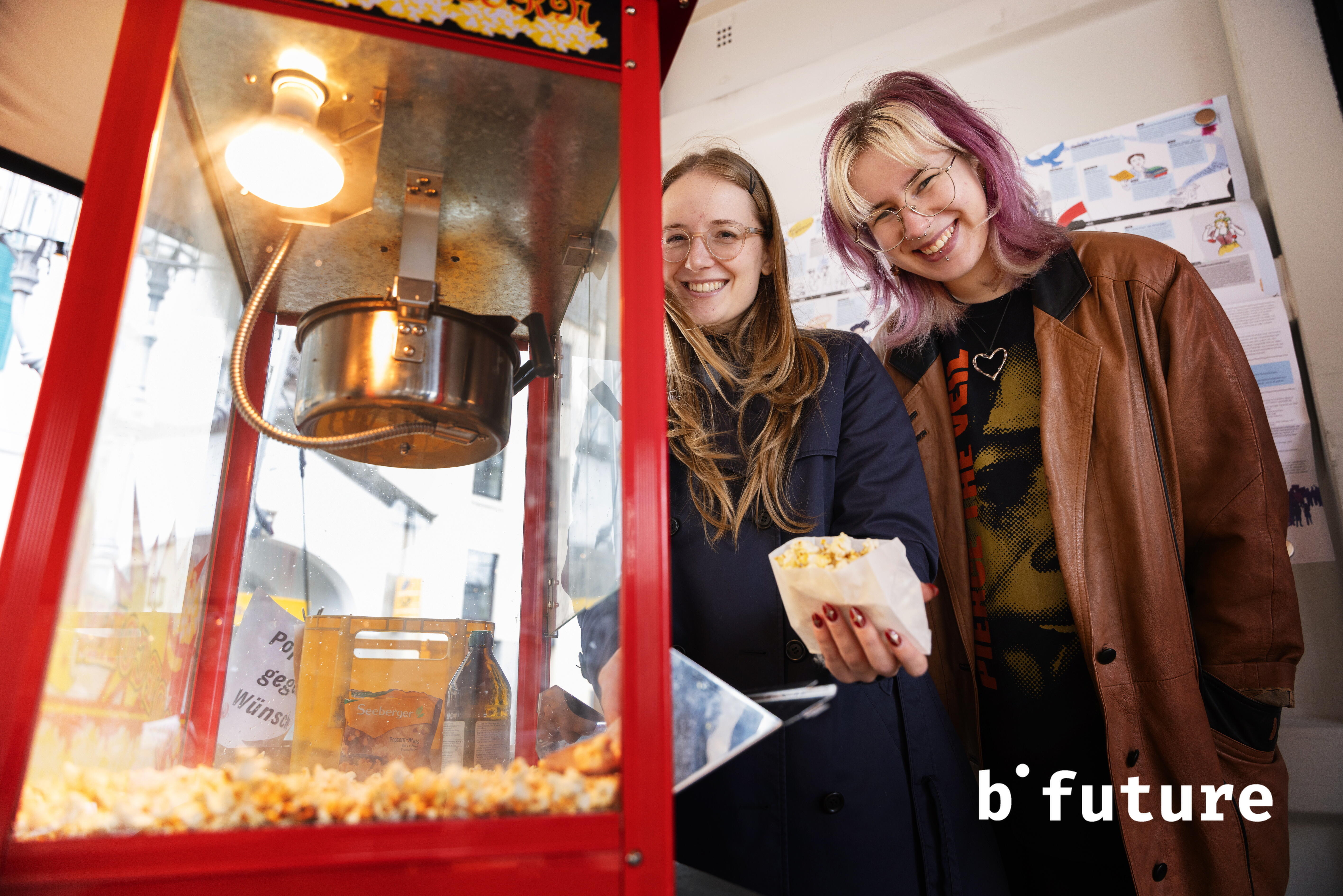 Zwei junge Personen stehen neben einer Popcorn-Maschine und lächeln in die Kamera