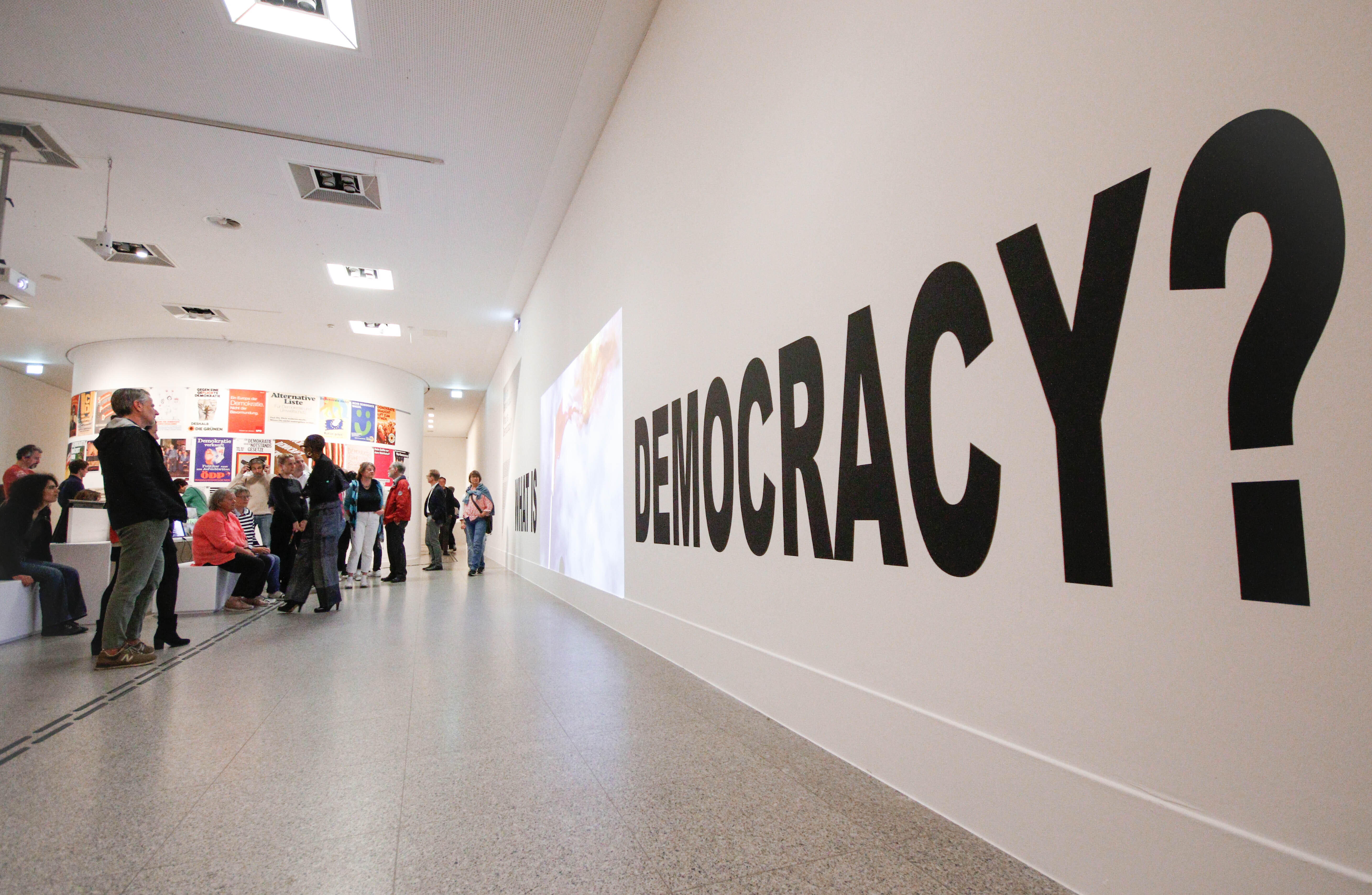Ein Bild aus der Demokratieausstellung der Bundeskunsthalle. Zusehen sind Menschen die vor einer Wand stehen, an der groß Democracy steht.