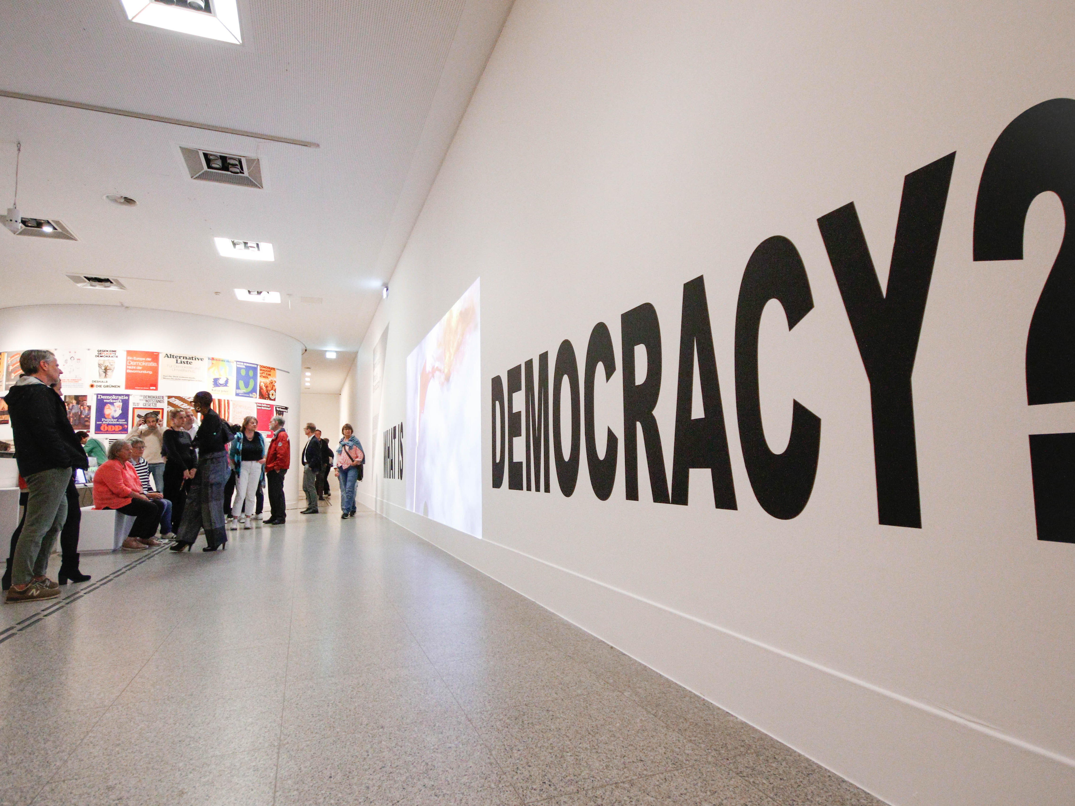 Ein Bild aus der Demokratieausstellung der Bundeskunsthalle. Zusehen sind Menschen die vor einer Wand stehen, an der groß Democracy steht.