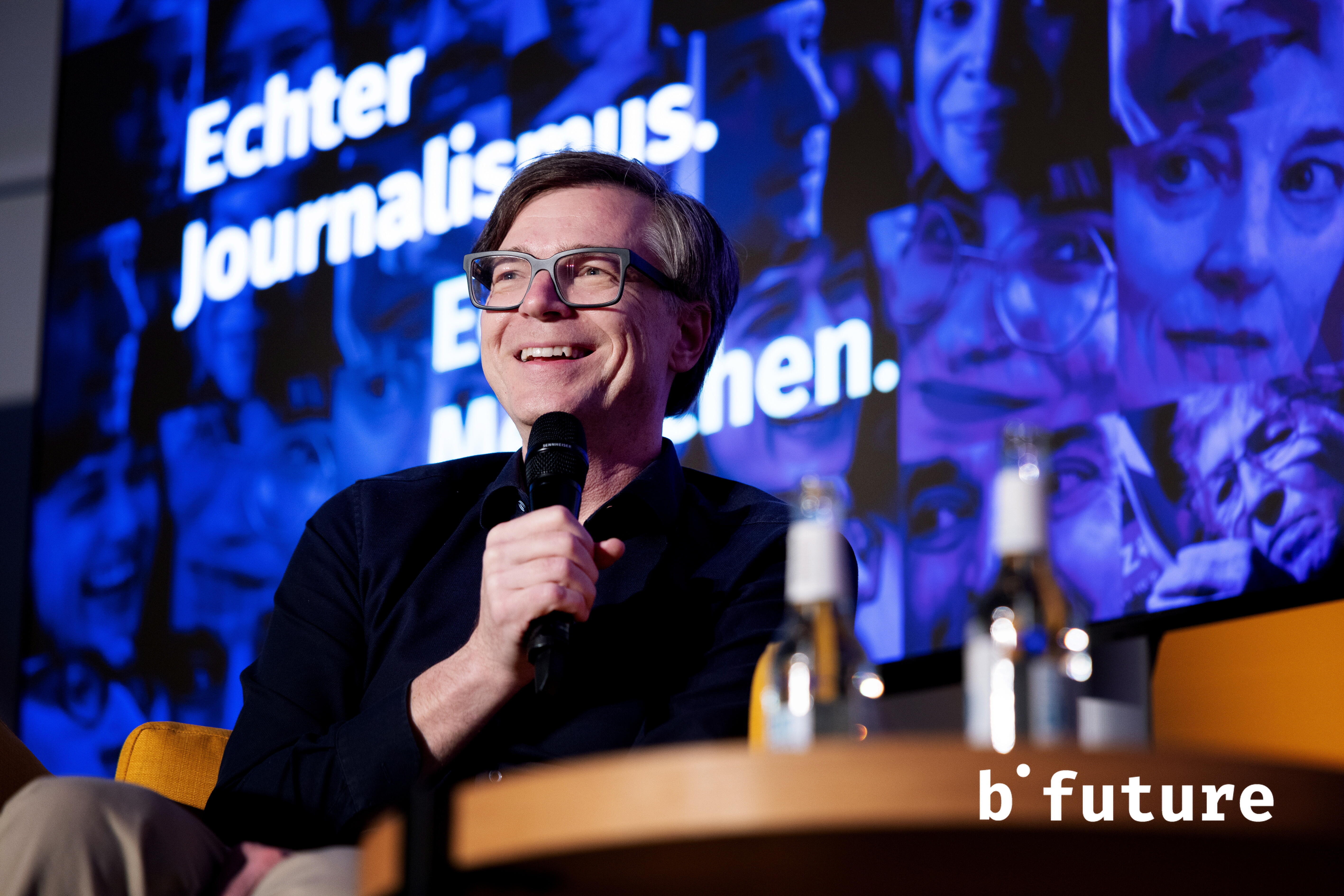 Ralph Caspers sitzt auf einer Bühne. Er hält ein Mikrofon in der Hand und lächelt.