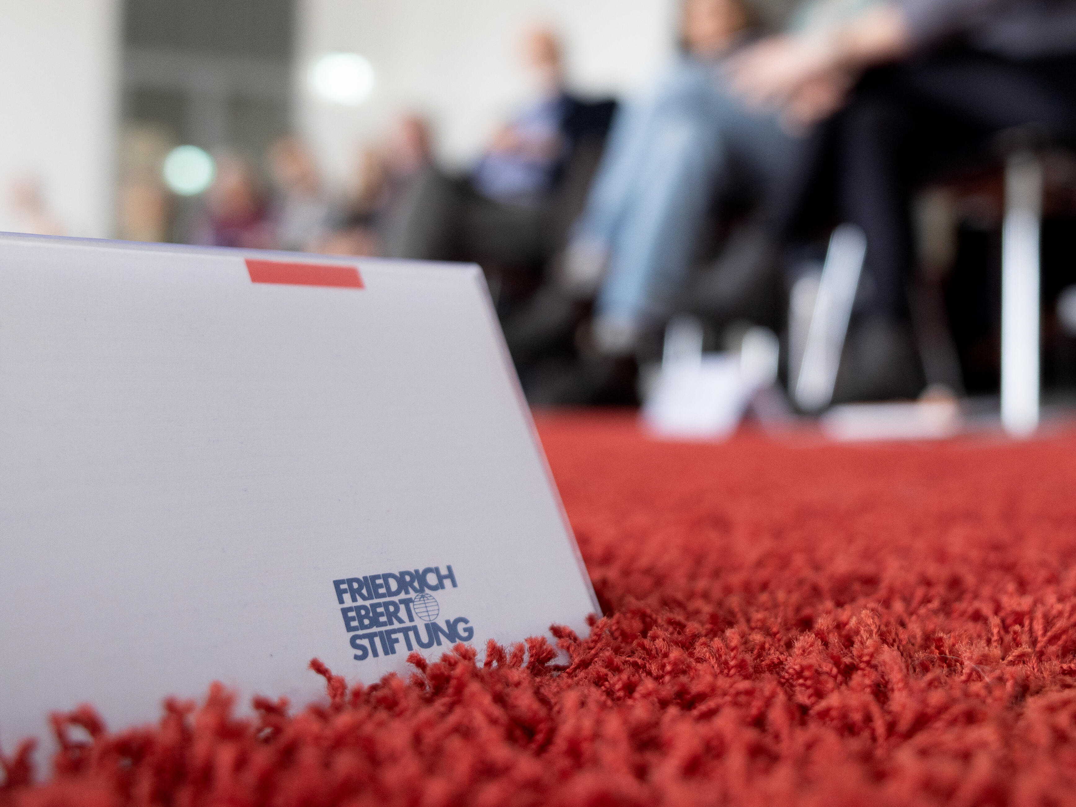 Ein umgedrehtes Namensschild mit dem Logo der Friedrich-Ebert-Stiftung steht auf einem Teppichboden. Im Hintergrund erkennt man in der Unschärfe sitzende Festivalbesuchende.