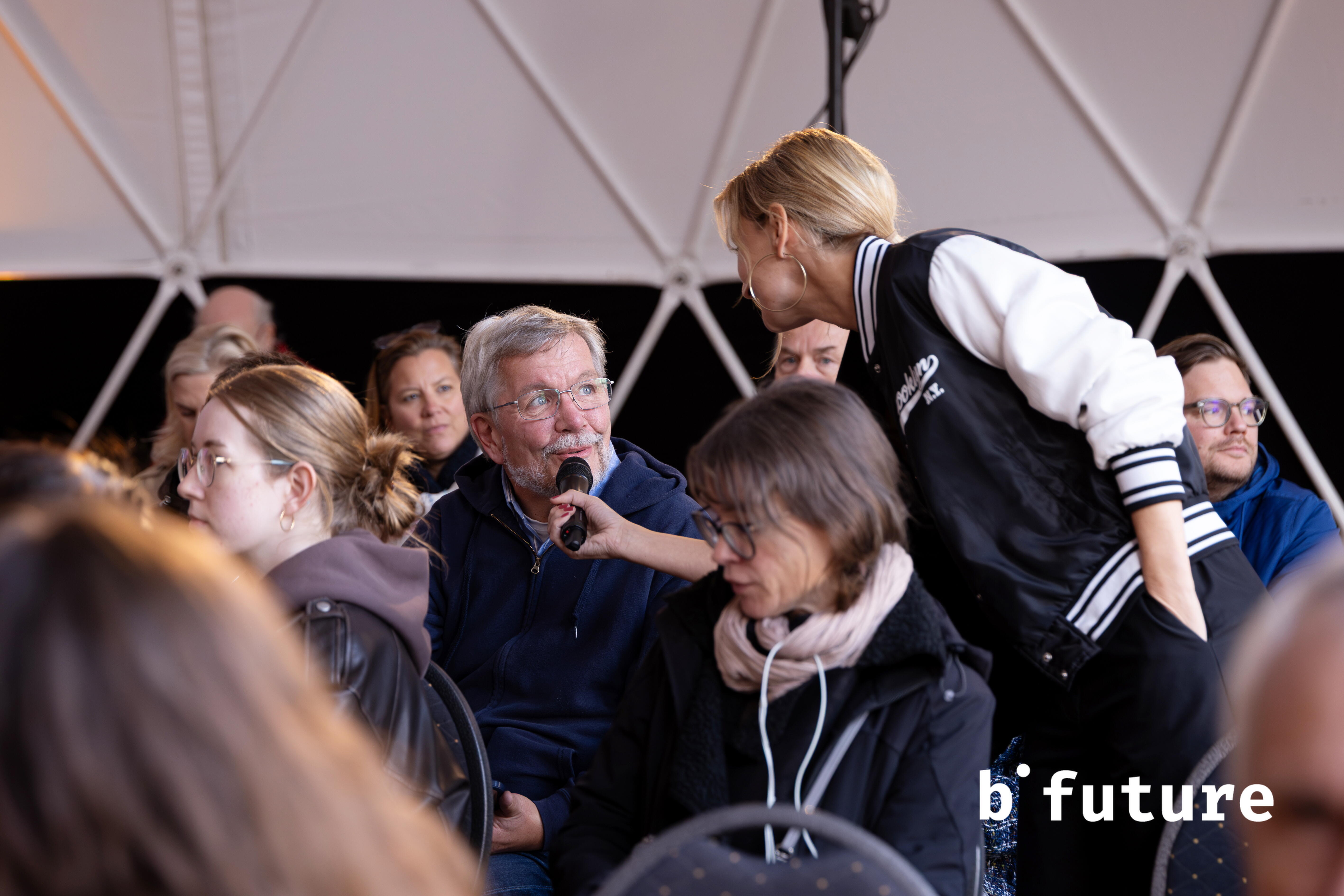 Ein Mann sitzt im Zuschauerraum, ihm wird von einer Frau ein Mikro an den Mund gehalten.
